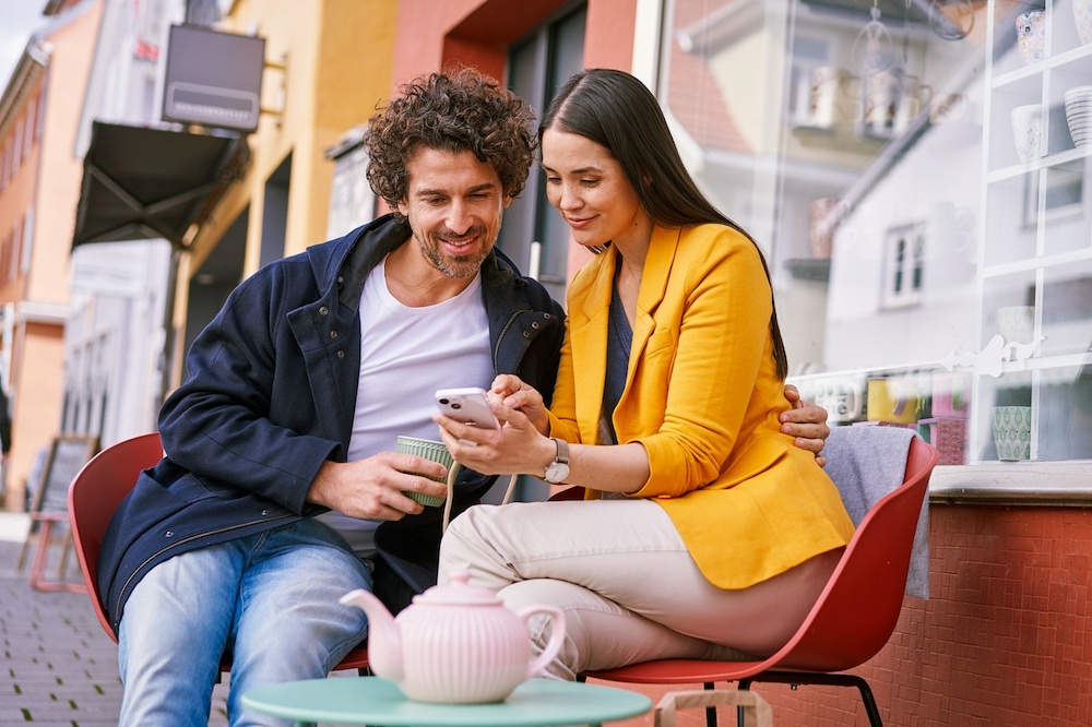 Ein Mann und eine Frau sitzen draußen an einem Tisch und sehen sich etwas auf einem Smartphone an.