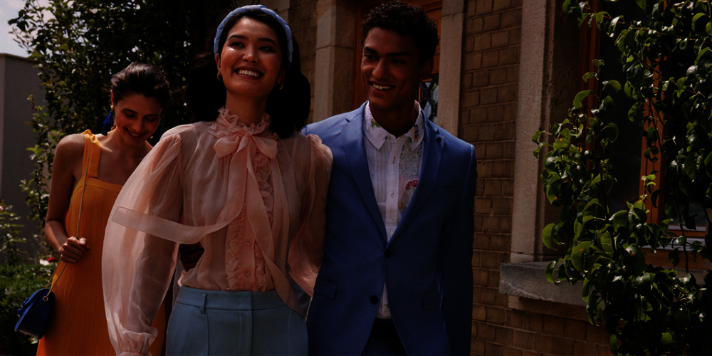 Drei Model in stylischer Kleidung. Frau mit rose Bluse und Haarreif. Mann mit weißen Hemd und Blazer im Hintergrund eine Frau mit einem gelben Kleid. 