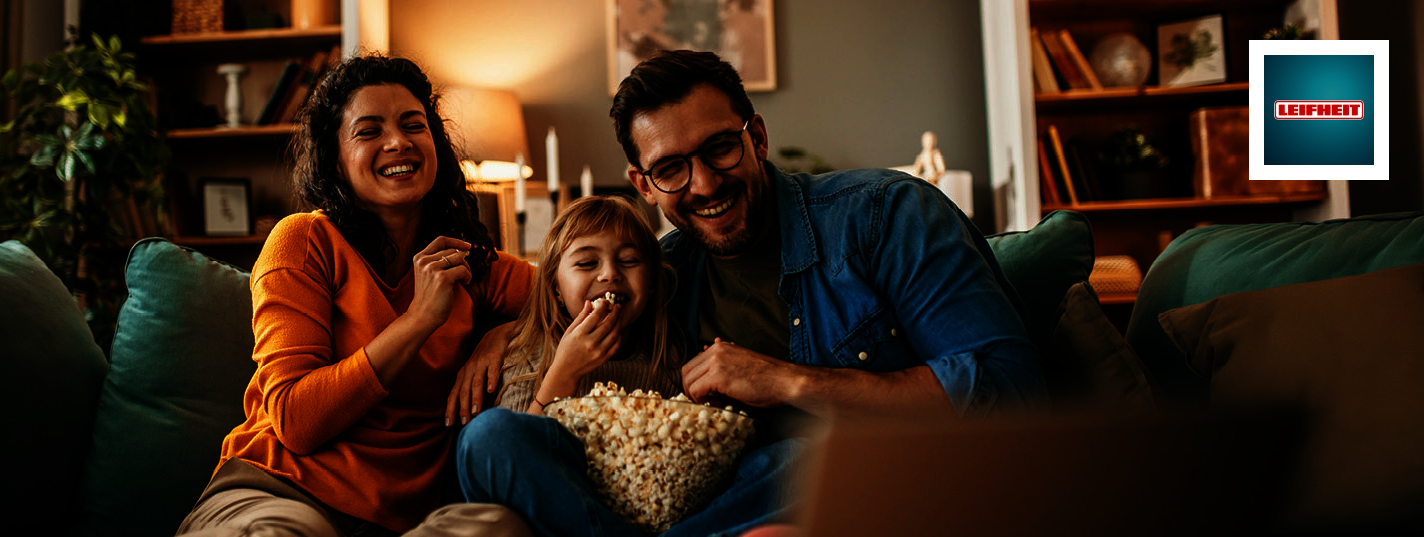 Drei Personen sitzen gemeinsam auf einem Sofa in einem Wohnzimmer und essen Popcorn aus einer großen Schüssel. Im Hintergrund sind Regale mit Büchern und Dekoration, Pflanzen sowie warme Lichtquellen zu sehen. Rechts oben im Bild ist das Leifheit-Logo ein