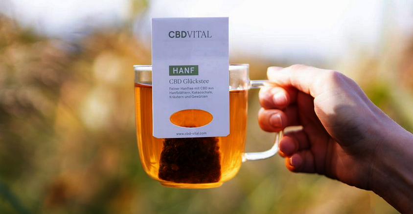 Close up von einer Hand die eine Tasse in der Hand hält mit einem Teebeutel auf dem Hanf steht