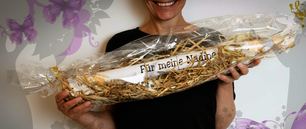 Ausschnitt einer Person, die ein persönliches Geschenk in den Händen hält. Durchsichtige Folie mit Stroh und einer Rolle mit einer Aufschrift "Für meine Nadine".