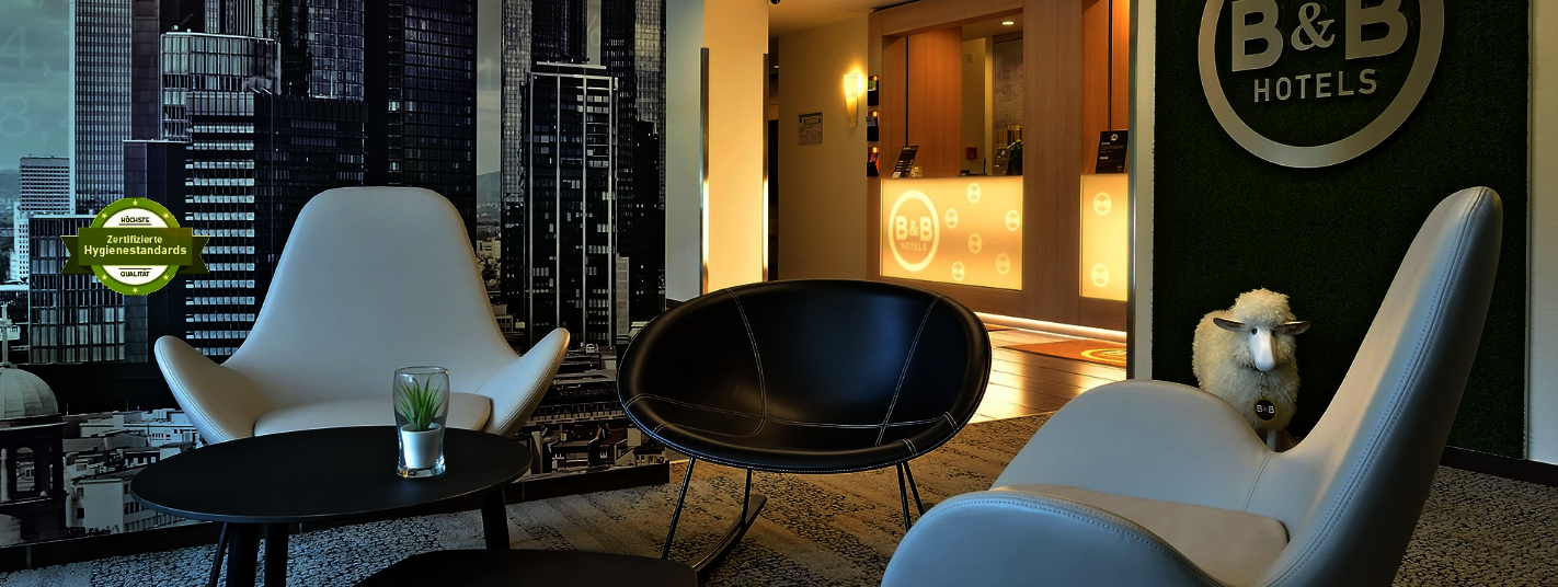 Lobbybereich eines B&B Hotels mit Sesseln, Tisch und Empfangstheke. Im Hintergrund sind das B&B Hotels-Logo sowie ein Hinweis auf zertifizierte Hygienestandards zu sehen.
