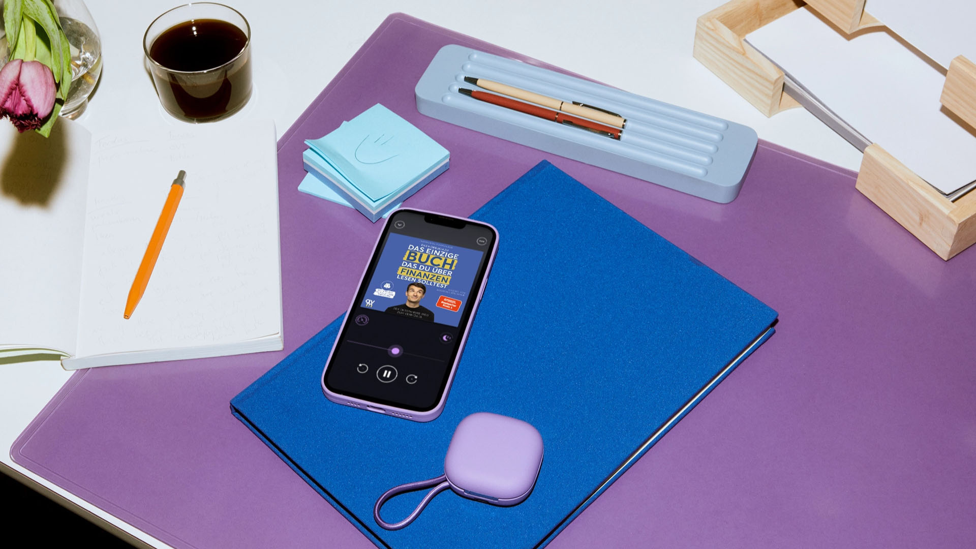 Smartphone mit geöffneter Hörbuch-App auf einem Schreibtisch mit Notizbuch, Stiften, Haftnotizen und einer Tasse Kaffee.